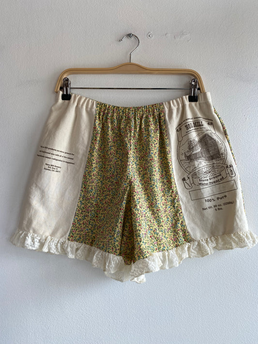 A pair of handmade shorts hanging from a wooden hanger against a white wall. The shorts feature a central panel of green, yellow, and orange floral print, flanked by cream-colored side panels with printed text and illustrations, finished with a ruffled lace hem.