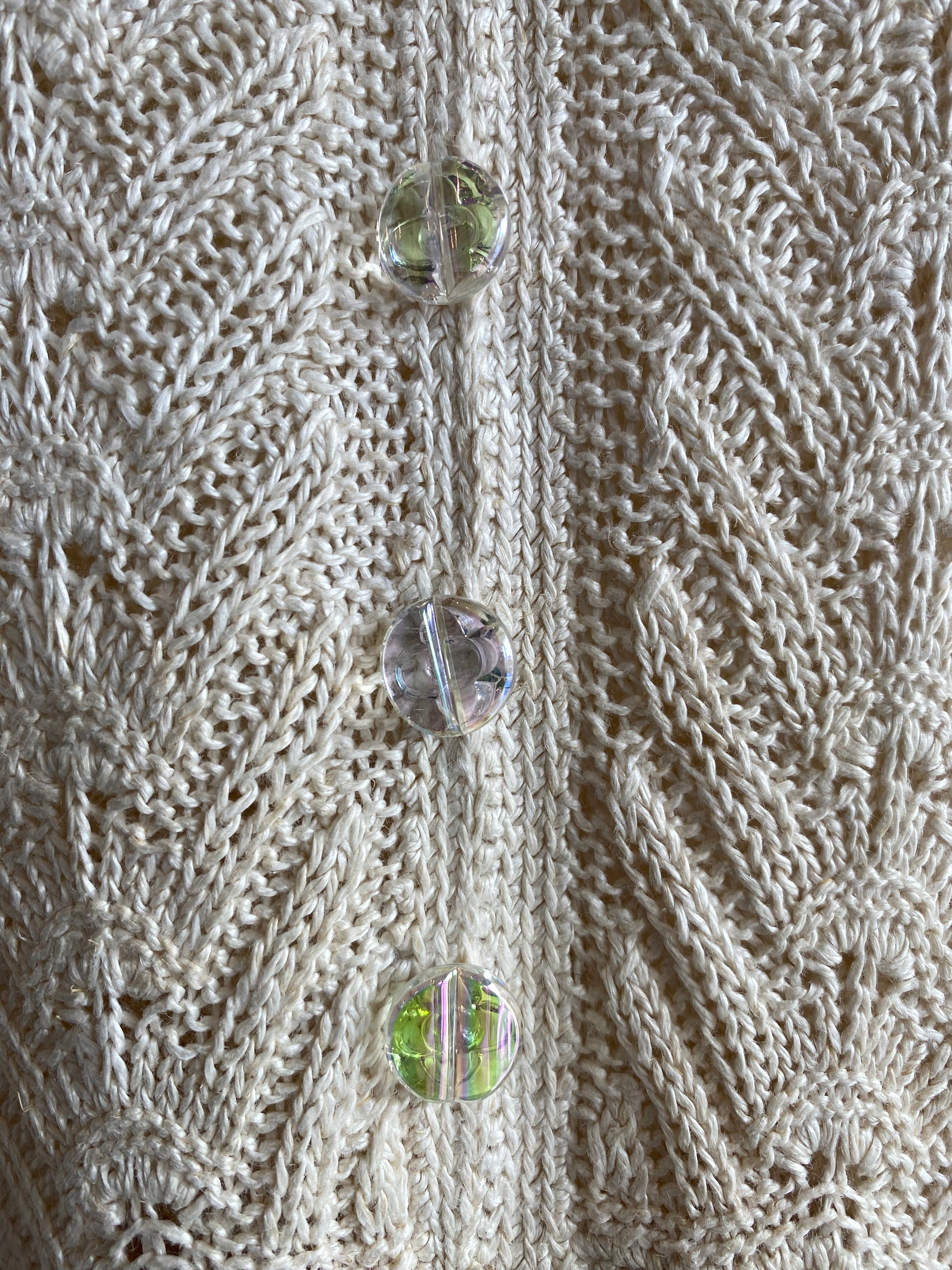 Close-up of a cream cable-knit sweater with three clear faceted buttons featuring iridescent green and pink reflections.