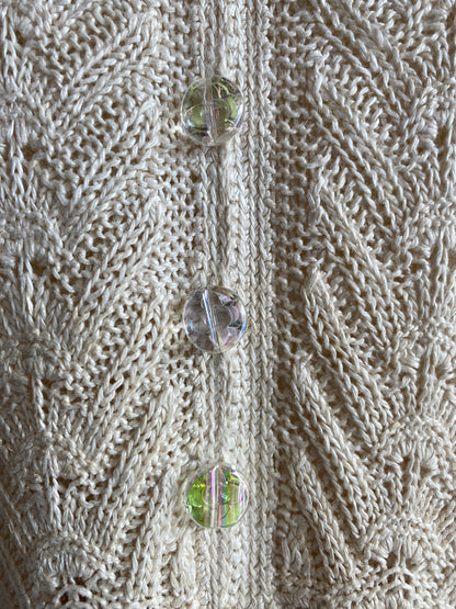 Close-up of a cream cable-knit sweater with three clear faceted buttons featuring iridescent green and pink reflections.