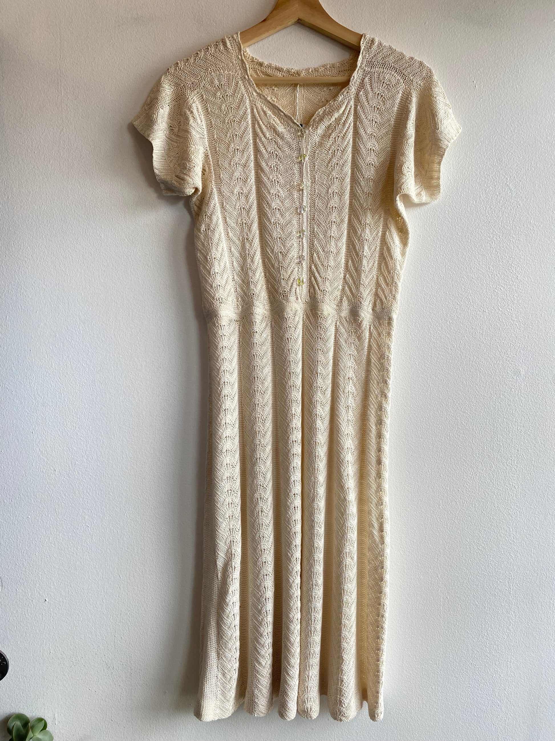 A cream-colored cable-knit short-sleeve dress hanging on a wooden hanger against a white wall.