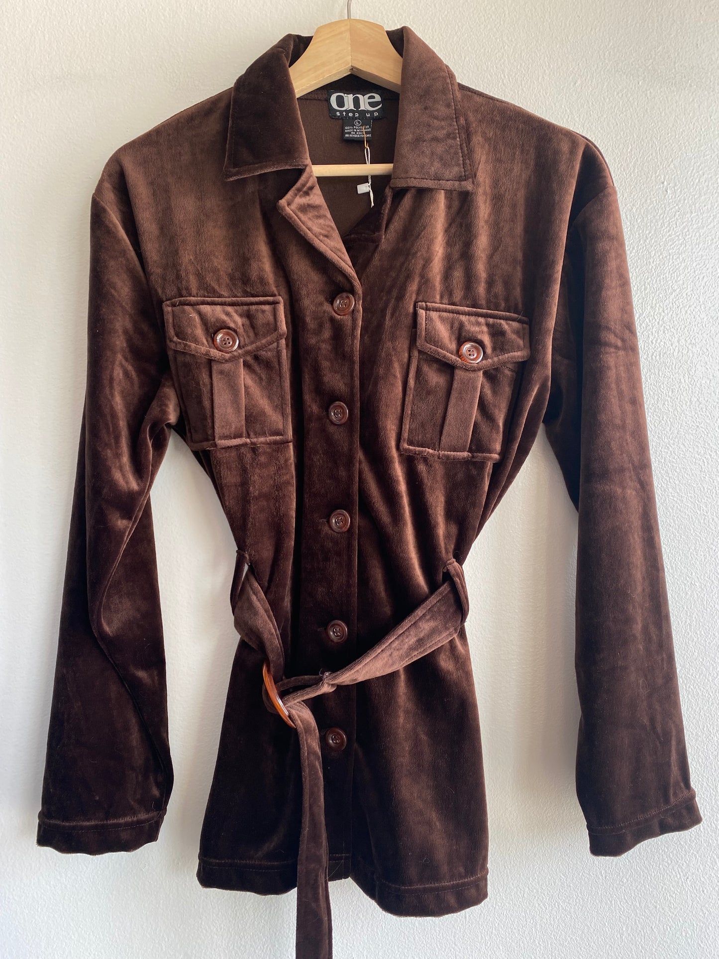 A dark brown velvet long-sleeve shirt jacket with a matching tie belt, hanging on a wooden hanger against a white wall. The brand label 'one step up' is visible inside the collar.