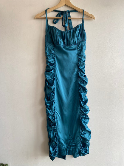 A teal silk dress with ruffle details hanging on a wooden hanger against a plain off-white wall.