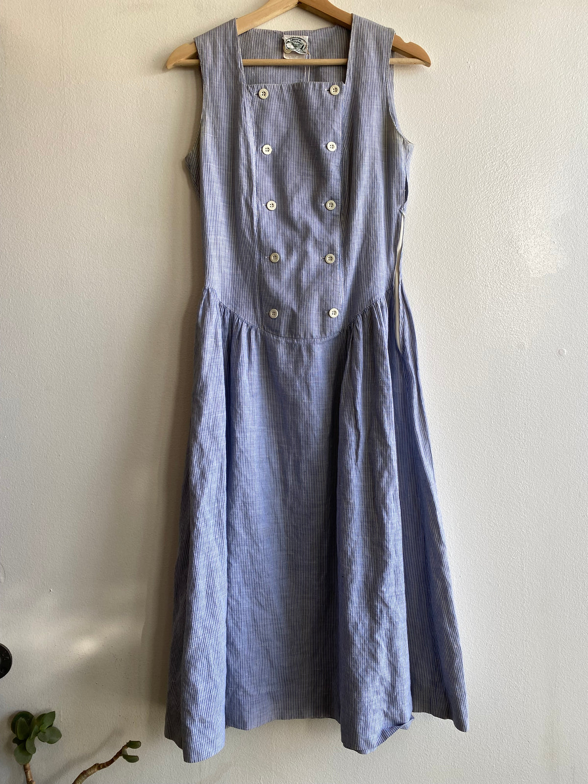 A sleeveless dusty blue linen dress with a double-breasted button front, hanging on a wooden hanger against a plain off-white wall.