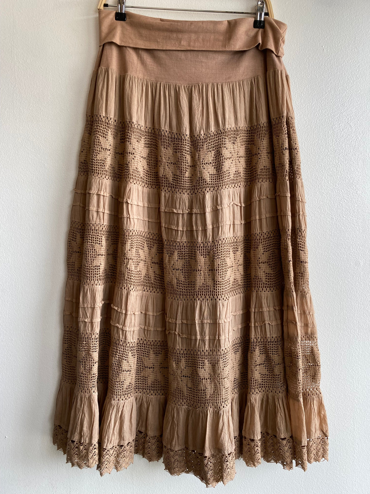 A tiered midi skirt in a warm taupe-brown color, featuring horizontal rows of embroidered geometric patterns and a matching fabric waistband, hanging against a white wall.