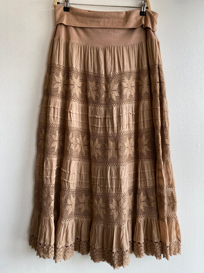 A tiered midi skirt in a warm taupe-brown color, featuring horizontal rows of embroidered geometric patterns and a matching fabric waistband, hanging against a white wall.