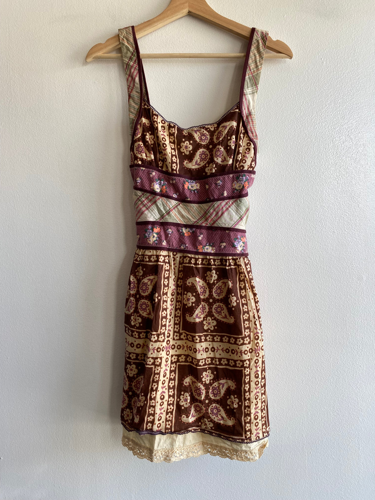 A sleeveless mini dress with a mixed patchwork print in brown, beige, and purple, hanging on a wooden hanger against a white wall.