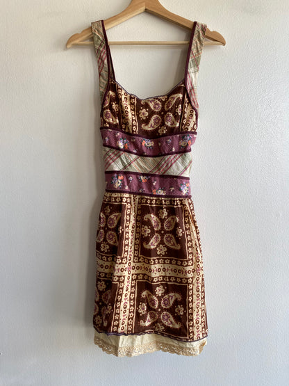A sleeveless mini dress with a mixed patchwork print in brown, beige, and purple, hanging on a wooden hanger against a white wall.