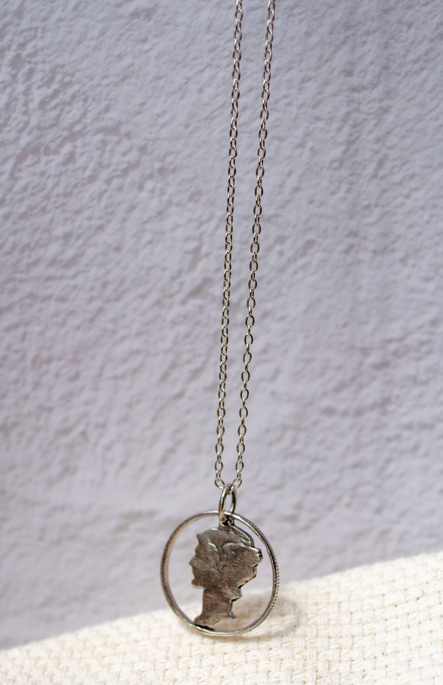 A silver chain necklace with a circular pendant featuring a profile silhouette of Roman god Mercury's face, displayed against a textured light background.