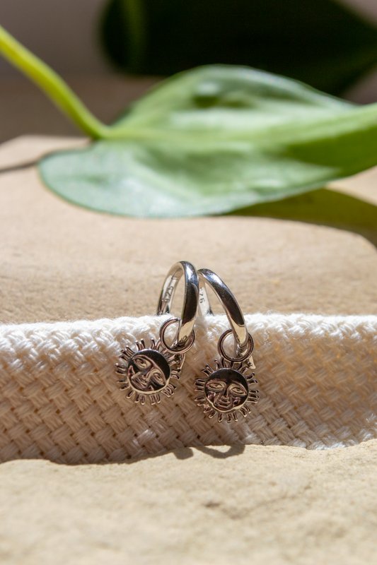 A pair of silver hoop earrings with sun and moon charms, displayed on a textured white woven band over a sandy surface with a green leaf in the background.